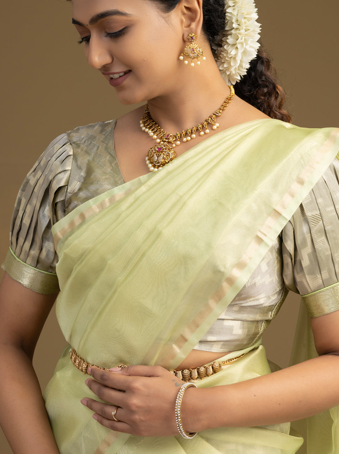 Woman wearing a light green saree with gold jewelry against a beige background
